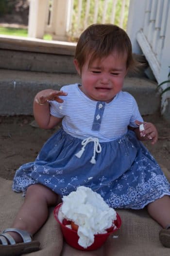 Toddler girl wearing blue dress crying while smashing cake 
