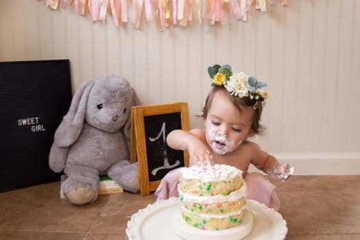 Toddler girl with flowers on head smashing a three tier cake with frosting and sprinkles while gray stuffed bunny watches in background