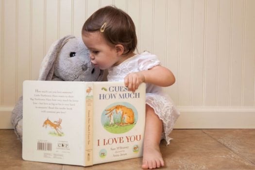 Toddler girl kissing gray bunny stuff animal reading Guess How Much I Love You book 