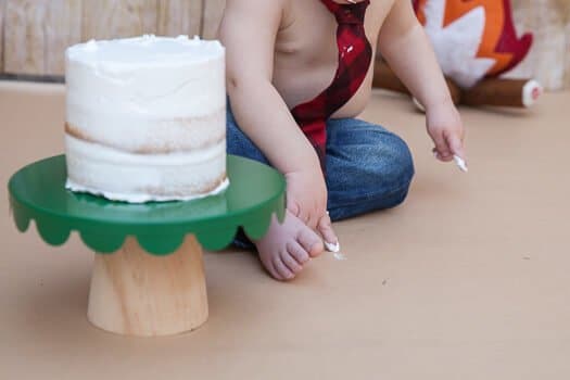 Three tier white cake on green pedestal with toddler wearing a tie and jeans 