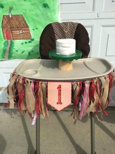 Three tier white cake on green pedestal on toddler chair with number 1 banner around it 