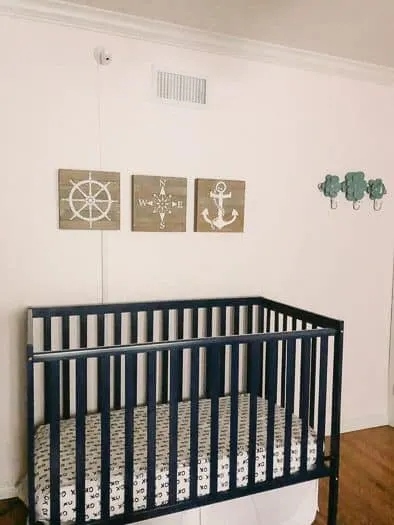 Slightly off-center view of a kids bedroom. Blue crib with three square wood pictures hanging above. Pictured on the wood squares are: the wheel of a ship, compass, and an anchor. 