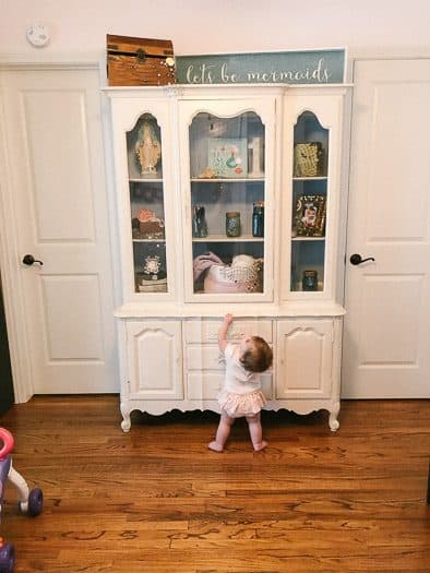 White hutch with let's be mermaid on top and a pirate chest. Multiple knickknacks inside the hutch. A baby is standing at the bottom looking up towards the top of the hutch.