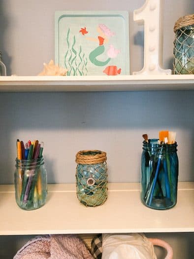 Blue tinted mason jars holding some art supplies, brushes and pencils.