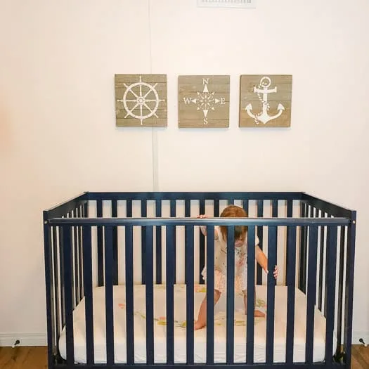 Wood wall art with a wheel of a ship, compass, and anchor, above a blue crib with a baby standing inside.