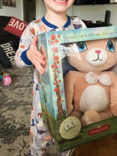Toddler wearing pajamas holding Binka Bear toy in package in living room 