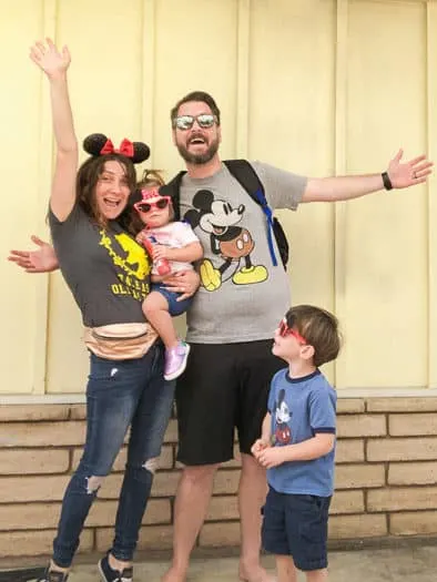 Family dressed as Disney Tourists. Mom in Belle shirt with Minnie ears and gold fanny pack, mom holding little girl with Minnie sunglasses, dad in Mickey shirt, little boy with Mickey sunglasses in a Mickey shirt