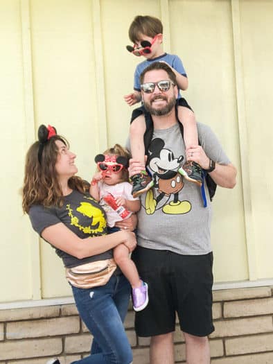 Family dressed as Disney Tourists. Mom in Belle shirt with Minnie ears, mom holding little girl with Minnie sunglasses, dad in Mickey shirt holding little boy on his head with Mickey sunglasses