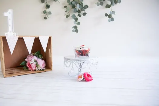 Eucalyptus leaves hanging in the background, a wooden box with flowers inside and a number one on top. A clear bowl holding blueberries and strawberries with whipped cream on a cake plate.