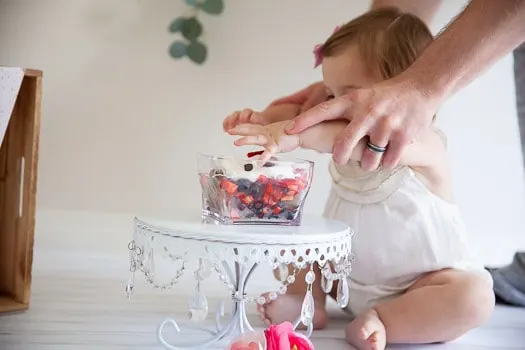 A little girl's hands being put into the clear bowl with strawberries and blueberries and whipped cream sitting on a cake plate