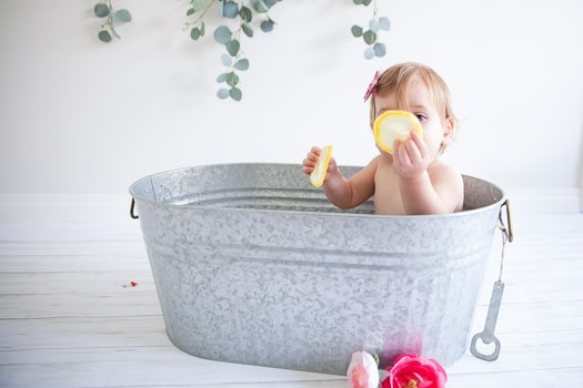 Little girl holding up a lemon slice in front of her face sitting in a galvanized tub with eucalyptus hanging in the background