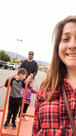 A man pushing a Home Depot wood cart with two kids ridding on it. A women in the foreground smiling.