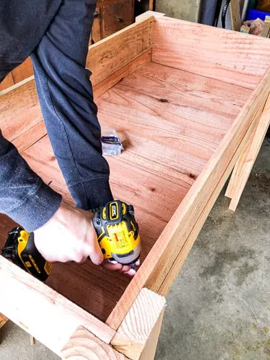 Inside of a raised garden bed showing a person holding a drill to screw in the slats on the bottom 