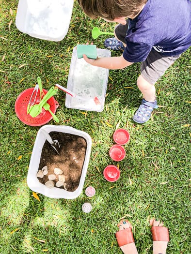 Container with mud and rocks, sitting next to a red colander with kitchen utensils sitting next to a bin of soapy water all sitting in the grass with a kid hovering above them.
