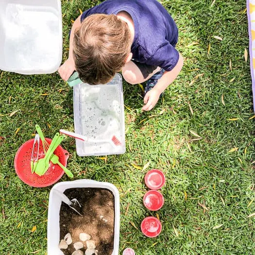 Kid playing in soapy water in a container with a colander with kitchen utensils sitting by and a bucket of dirt and rocks.