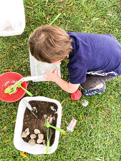 Kid playing in soapy water in a container with a colander with kitchen utensils sitting by and a bucket of dirt and rocks.