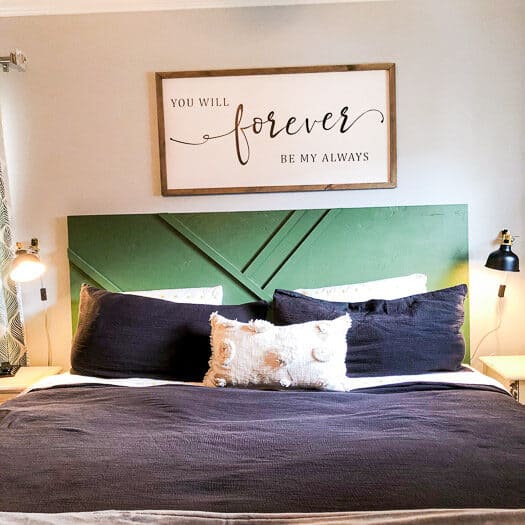A bed with black bedding, white throw pillows, a green geometric headboard and a wood frame above the bed.