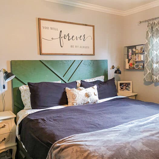 A bed with black bedding, white throw pillows, a green geometric headboard and a wood frame above the bed. To the side you can see black hanging lamps and a shelving unit.