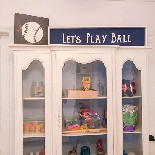 Curio cabinet filled with art supplies with a baseball and lets play ball artwork on top.