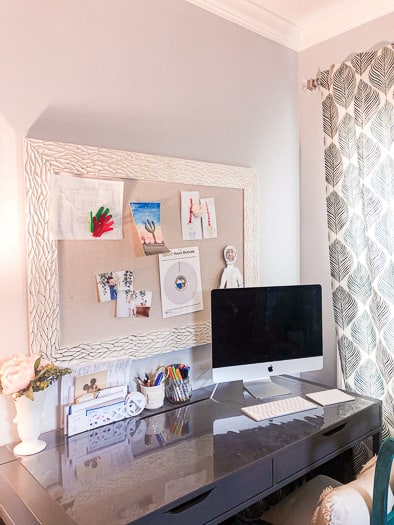 A desk holding a computer and pens, a mail holder, and flowers with a bulletin board over the desk.