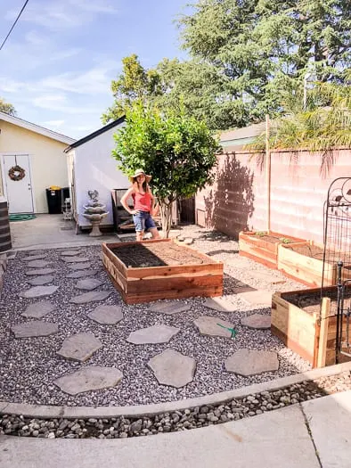 garden beds with dirt, rock and stepping stones around garden beds, women with pink shirt and flopping hat standing behind the garden bed