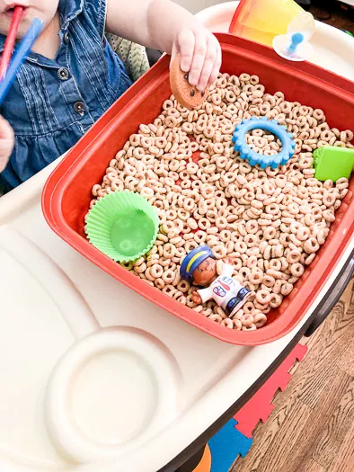 Cheerios in a red open container with toys inside and a small child reaching in.