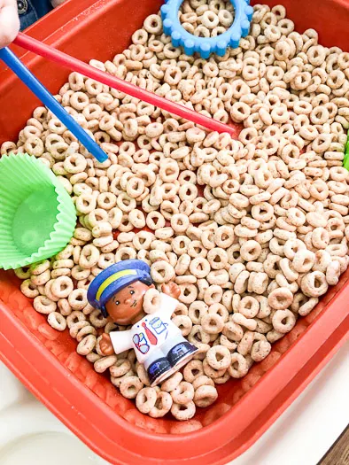 Cheerios in a red open container with toys inside and a small poking straws into the cheerios.
