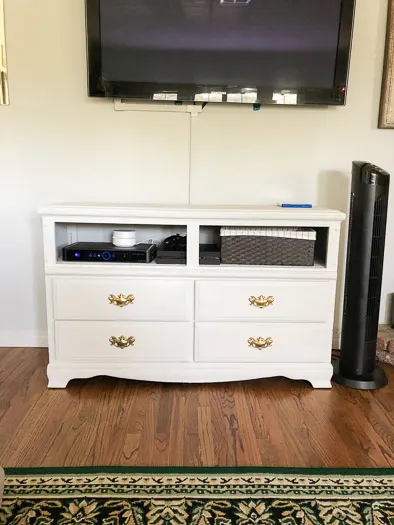 A white dresser with the top 2 drawers taken out and TV boxes in the opening and a basket. Gold handles on the bottom 4 drawers.