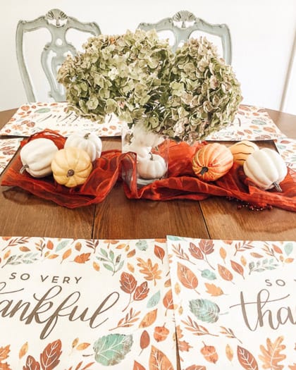 red table runner scrunched in center of table, clear vase with white pumpkins inside and hydrangeas coming out the top, pumpkins scattered around.