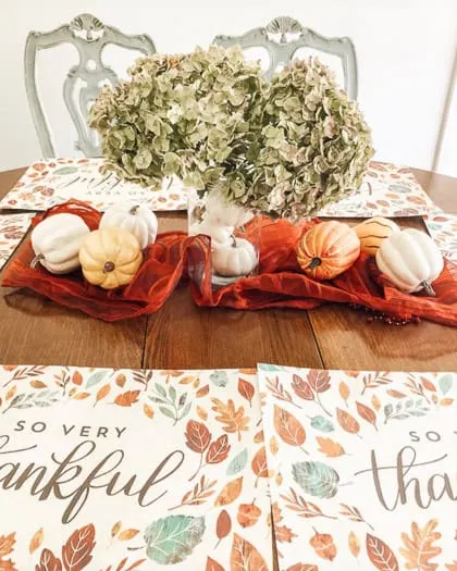 red table runner scrunched in center of table, clear vase with white pumpkins inside and hydrangeas coming out the top, pumpkins scattered around.