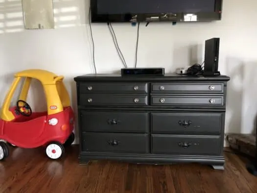 Black colored dresser with tv hangng on wall above and red toy car next to it