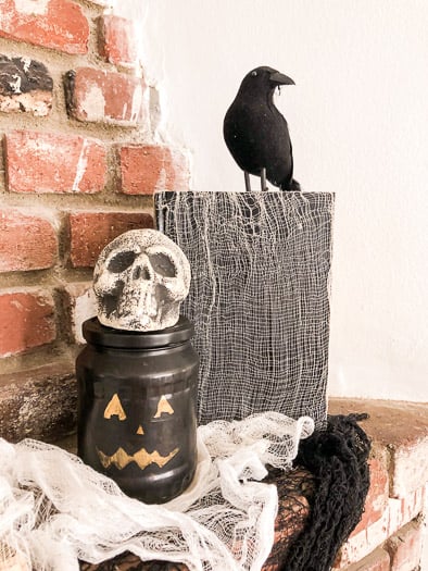 A cheese cloth covered book with a fake crow sitting on top, next to pumpkin jar with a skull on top
