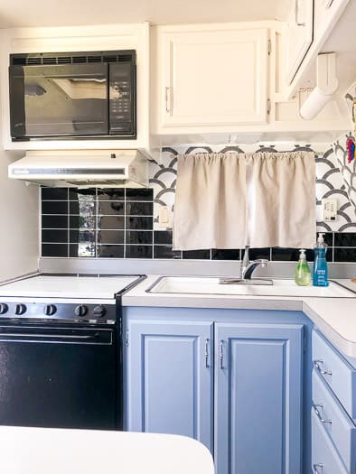 Kitchen view of a trailer with blue lower cabinets, white upper cabinets, scalloped wallpaper, and black subway tile backsplash.