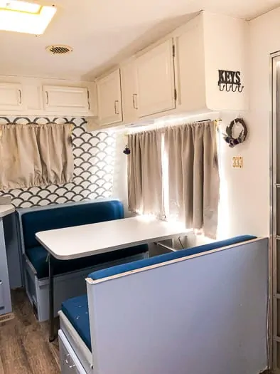 Dining table view of a trailer with blue cushions on dinette, blue lower cabinets, white upper cabinets, and scalloped wallpaper.