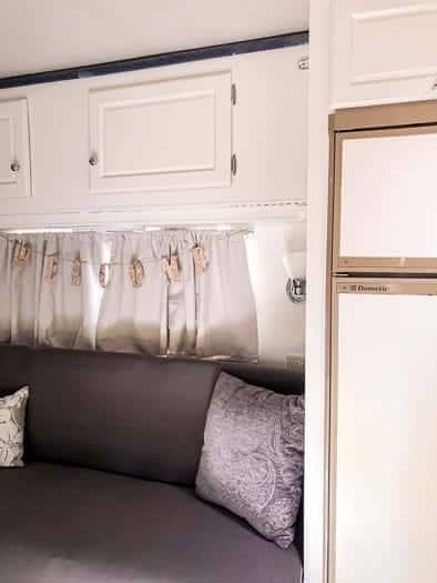 Grey couch with grey pillows a window over the couch with a hanging welcome sign, white upper cabinets in view and a fridge to the right side