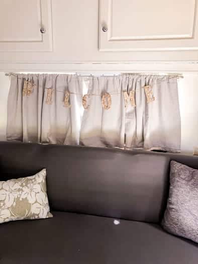 Grey couch with grey pillows a window over the couch with a hanging welcome sign