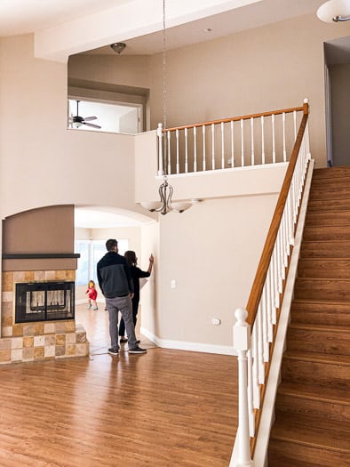 Two people walking through an empty house. Stairs on the right and a fireplace in the middle left