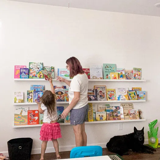 little girl reaching to put a book on the highest shelf with an adult standing near by