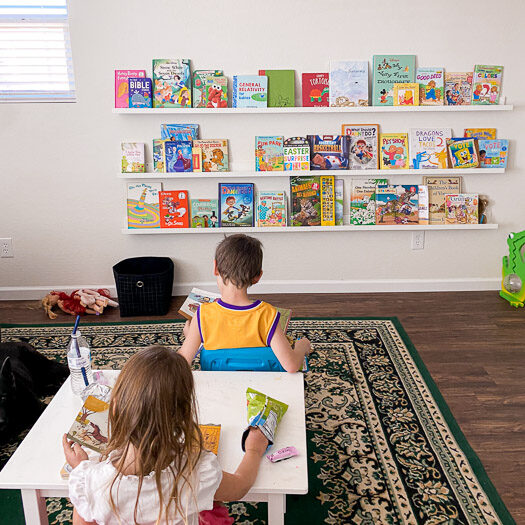 2 kids sitting at a table reading books in front of a wall with three long shelves holding books.