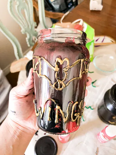 A jar with red and black marbled paint inside. The outside has a bat and the word wings made out of hot glue. The word and bat are painted gold.