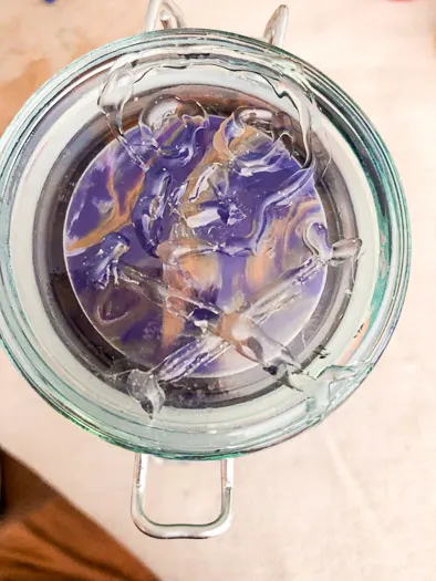 Top of a jar with purple and gold paint swirled inside. There is a clear skull made with hot glue on the top.