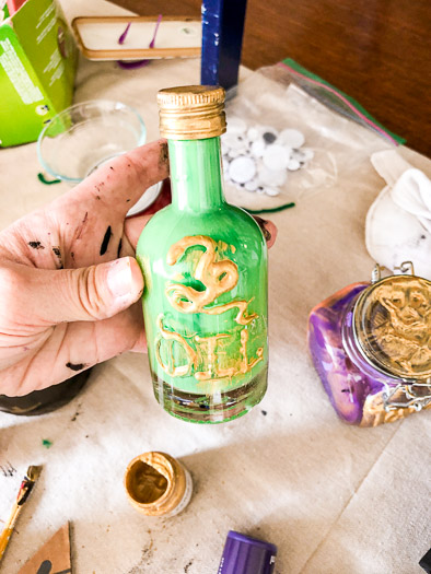 Clear bottle with green paint inside. Outside says the word oil and has the image of a snake made with hot glue. Both the word and snake are painted gold.