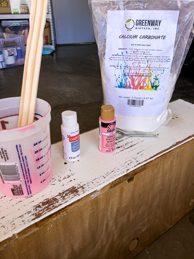 bag of calcium carbonate, 2 craft paint bottles - 1 pink 1 white, and a paint cup with pink paint and a stir stick sitting on a dresser drawer