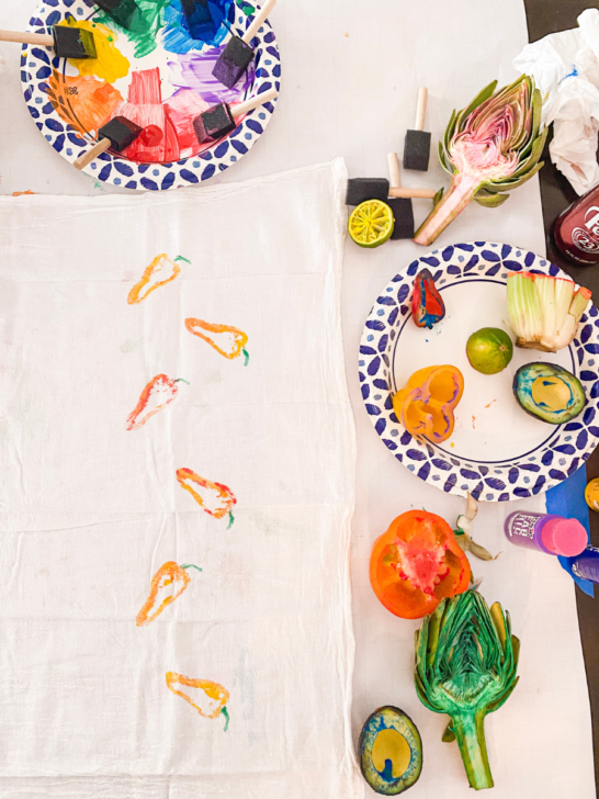 Veggies and fruit on the right side of the photo with paint on them. On the left side a towel with painted peppers.