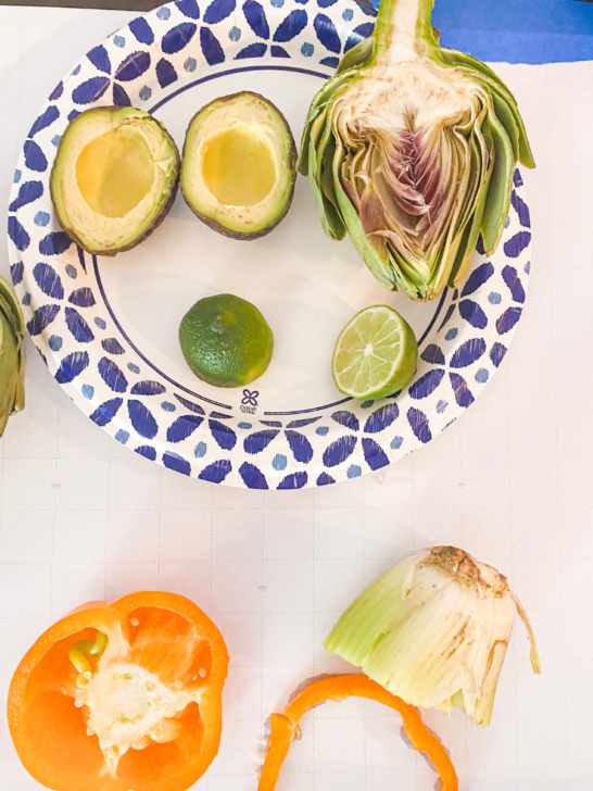 Artichoke, avocado, and limes sitting on a plate. Celery and bell pepper sitting near by.