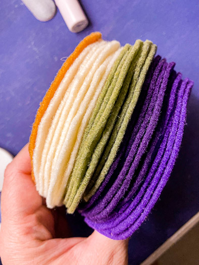 A hand holding a stack of felt circles orange on top, white, green, and purple on bottom.
