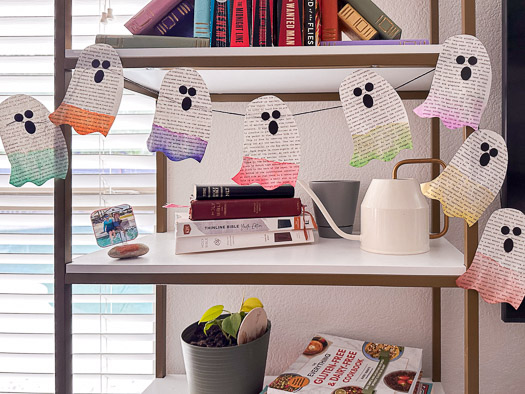 8 book page ghosts with all different colored bottom halves hanging on a bookshelf that has a stack of books and a watering can