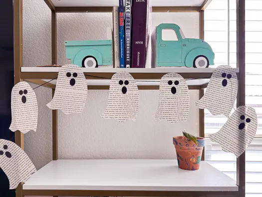 7 book page ghosts hanging on a bookshelf that has car bookends with books in between above it and a plant below it.